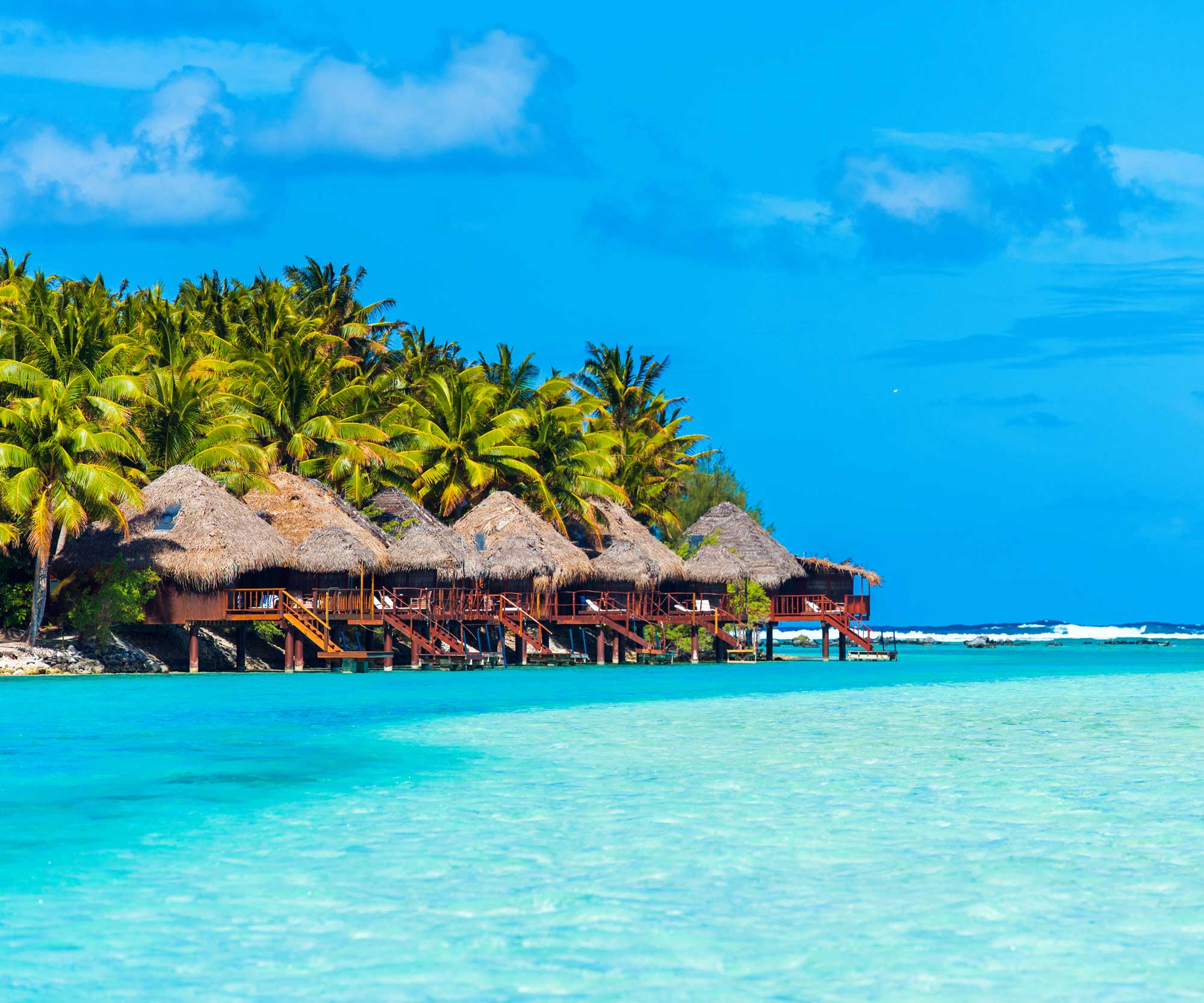 thatched houses sitting out over a lagoon with palm trees behind
